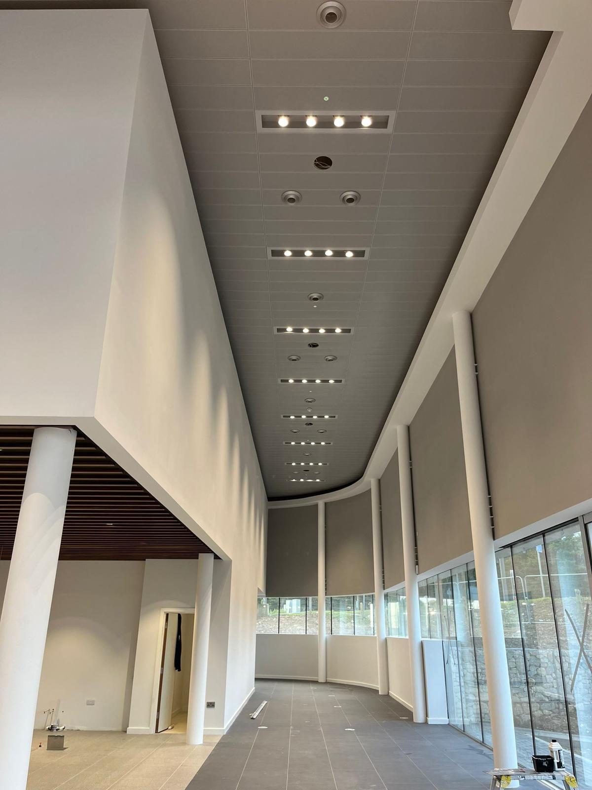 Curved plasterboard bulkhead edging a linear tile ceiling with inset lighting along a glazed corridor
