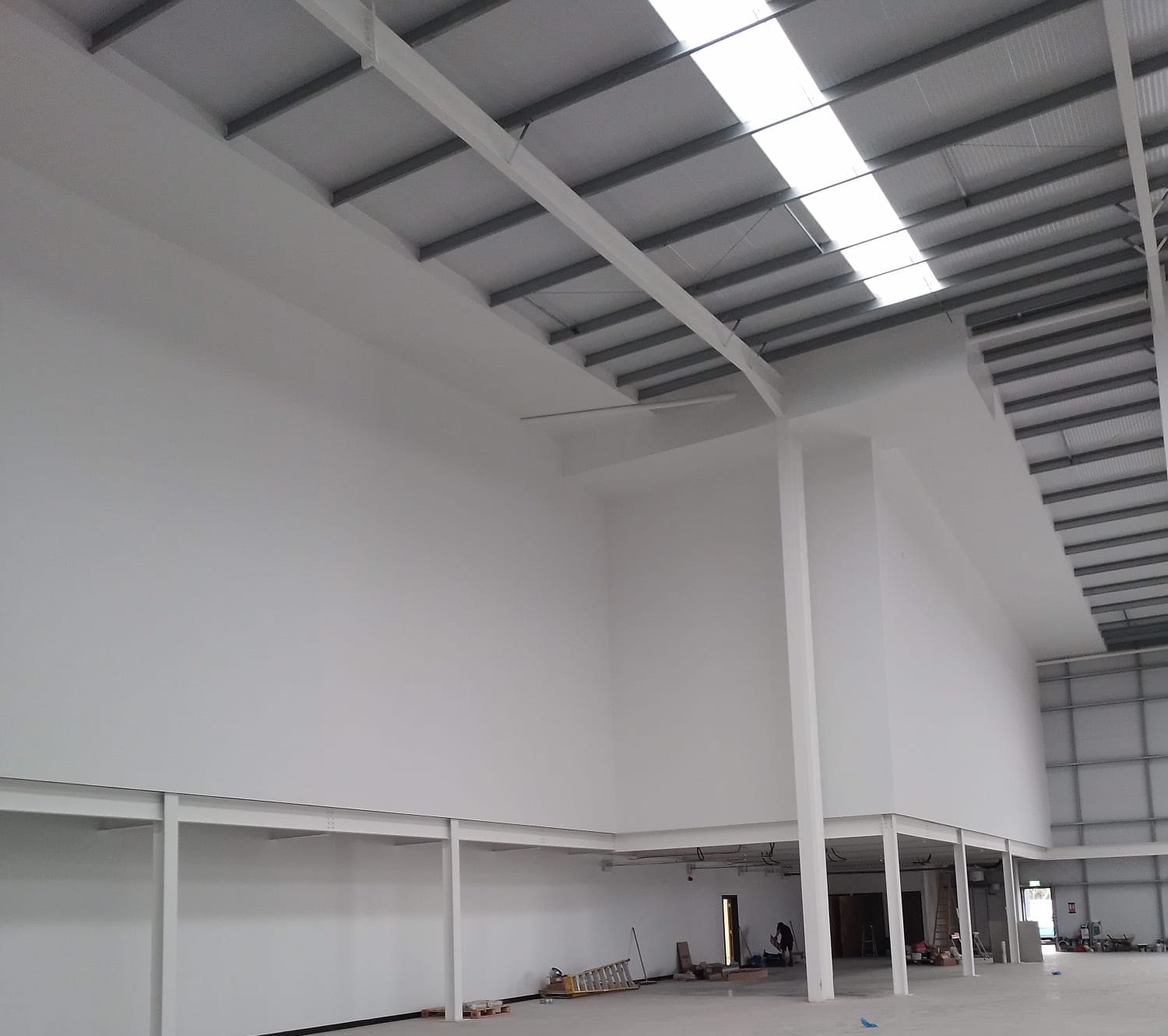 Full-height walls finished in white up to the roof steels of a warehouse, rising above a mezzanine